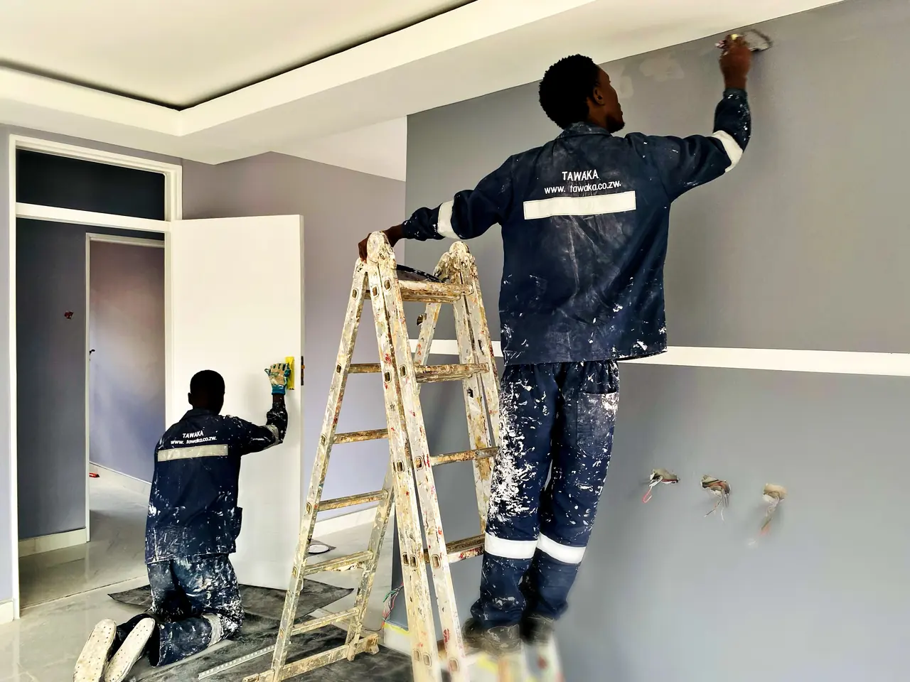 Two Tawaka Contracting painters in branded workwear finishing a grey feature wall and white reveals in Westgate, Zimbabwe